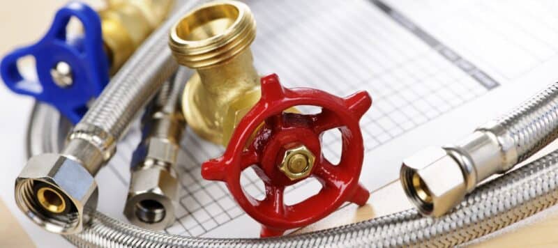 Close-up of various plumbing components, including a red valve handle, brass fittings, and flexible hoses, displayed on top of a maintenance checklist