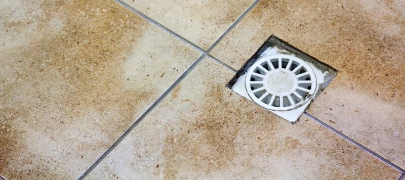 a white drain installed in under tan tile with a square cutout for access