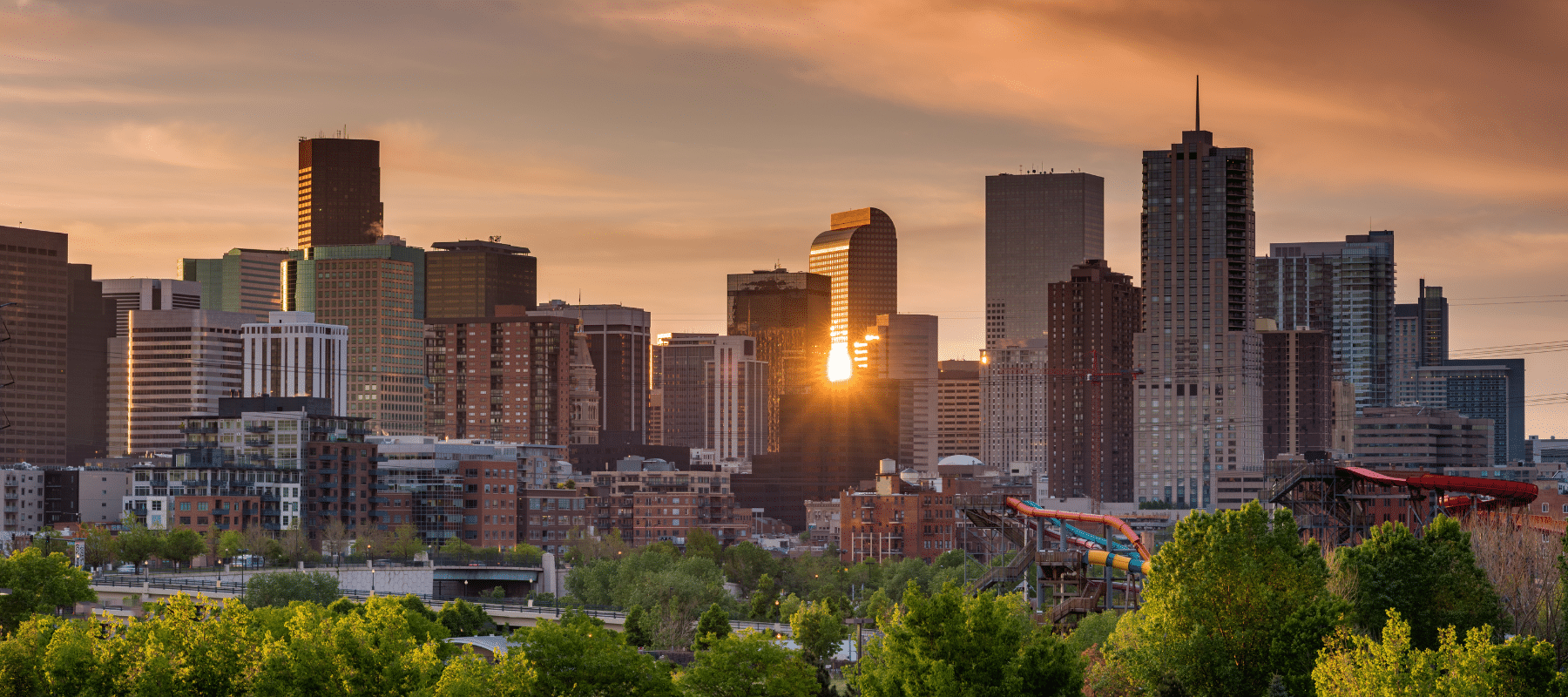 The Denver skyline at sunset, with warm sunlight reflecting off the glass buildings and a blend of urban structures surrounded by lush greenery